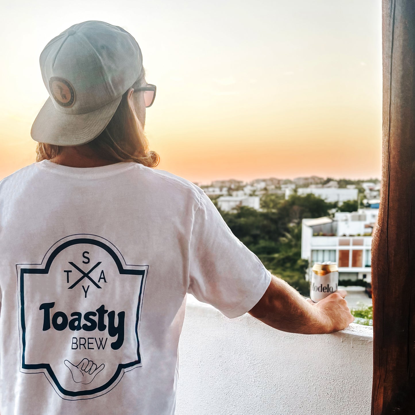 Man wearing white graphic tee with Toasty Brew beer label graphic back print with shaka hand graphic, rooftop view at sunset with a beer