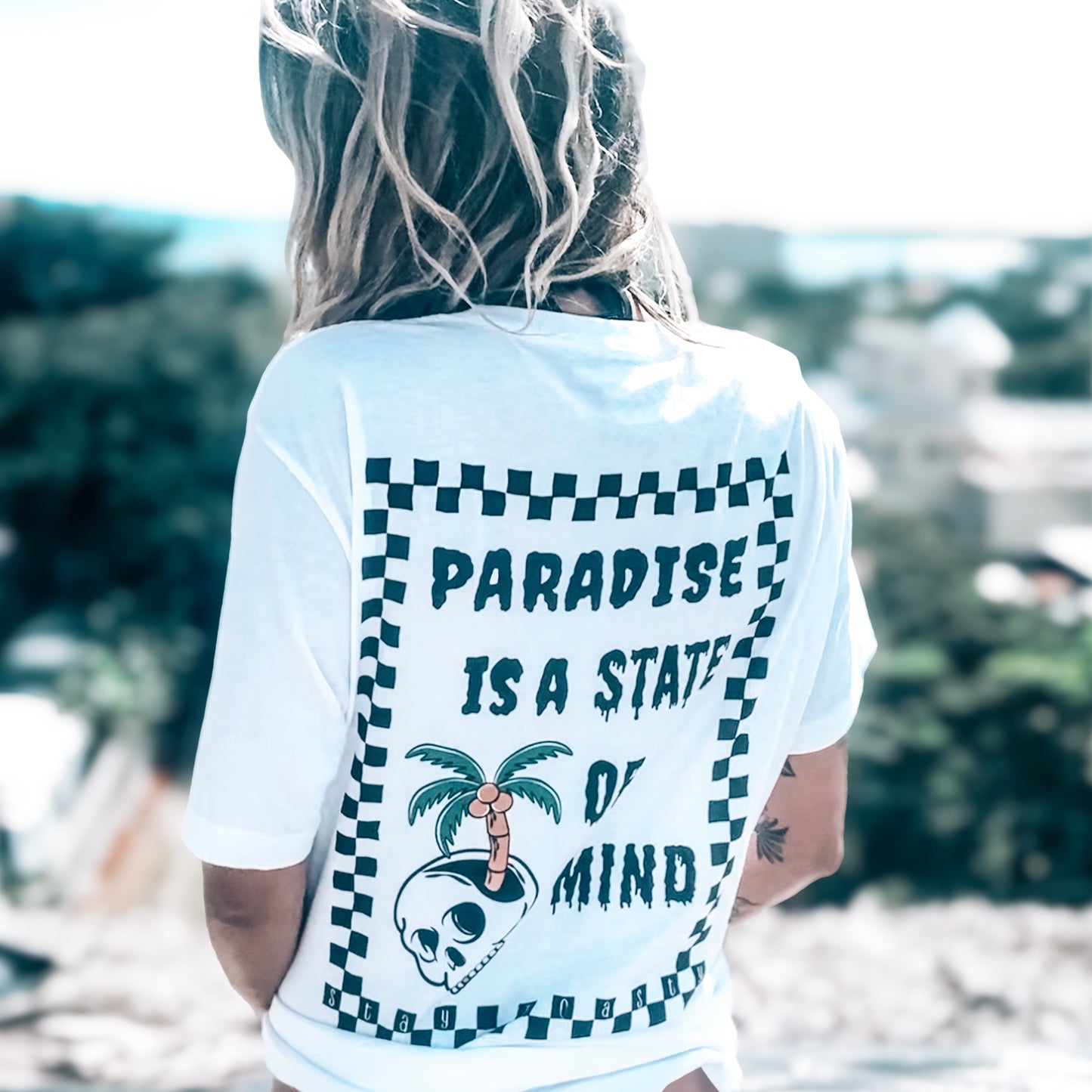 Person wearing a white t-shirt from the back view. The shirt has a palm tree skull design and text that says "Paradise is a state of mind" . The model is overlooking the ocean from a balcony