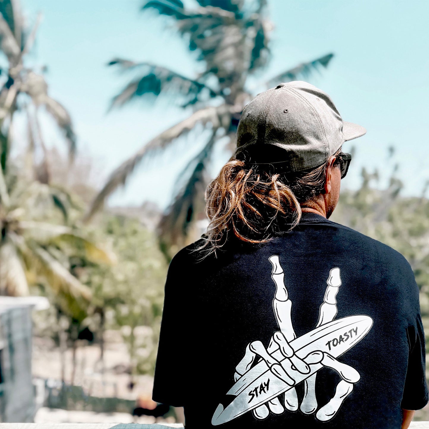 Black shirt with skeleton peace sign holding surfboard, worn by person overlooking tropical scene.