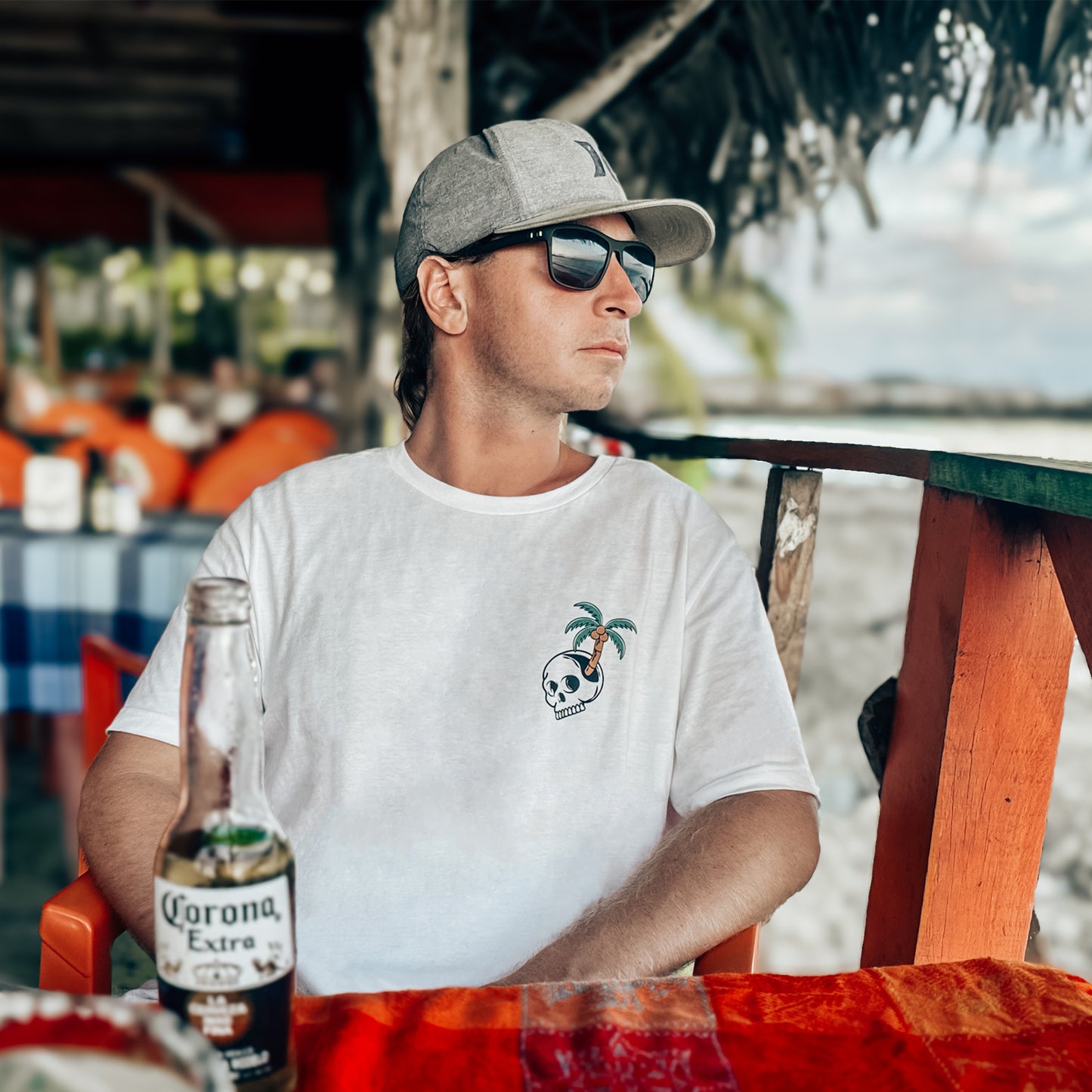 Male model sitting at beach bar with a corona beer. Wearing the Paradise State of Mind tee by Stay Toasty. Displays front chest graphic of a skull and palm tree