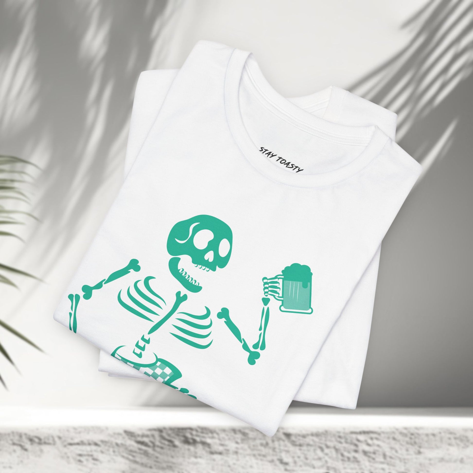 White t-shirt with a skeleton surfer design holding a beer and 'Stay Toasty' text shown folded