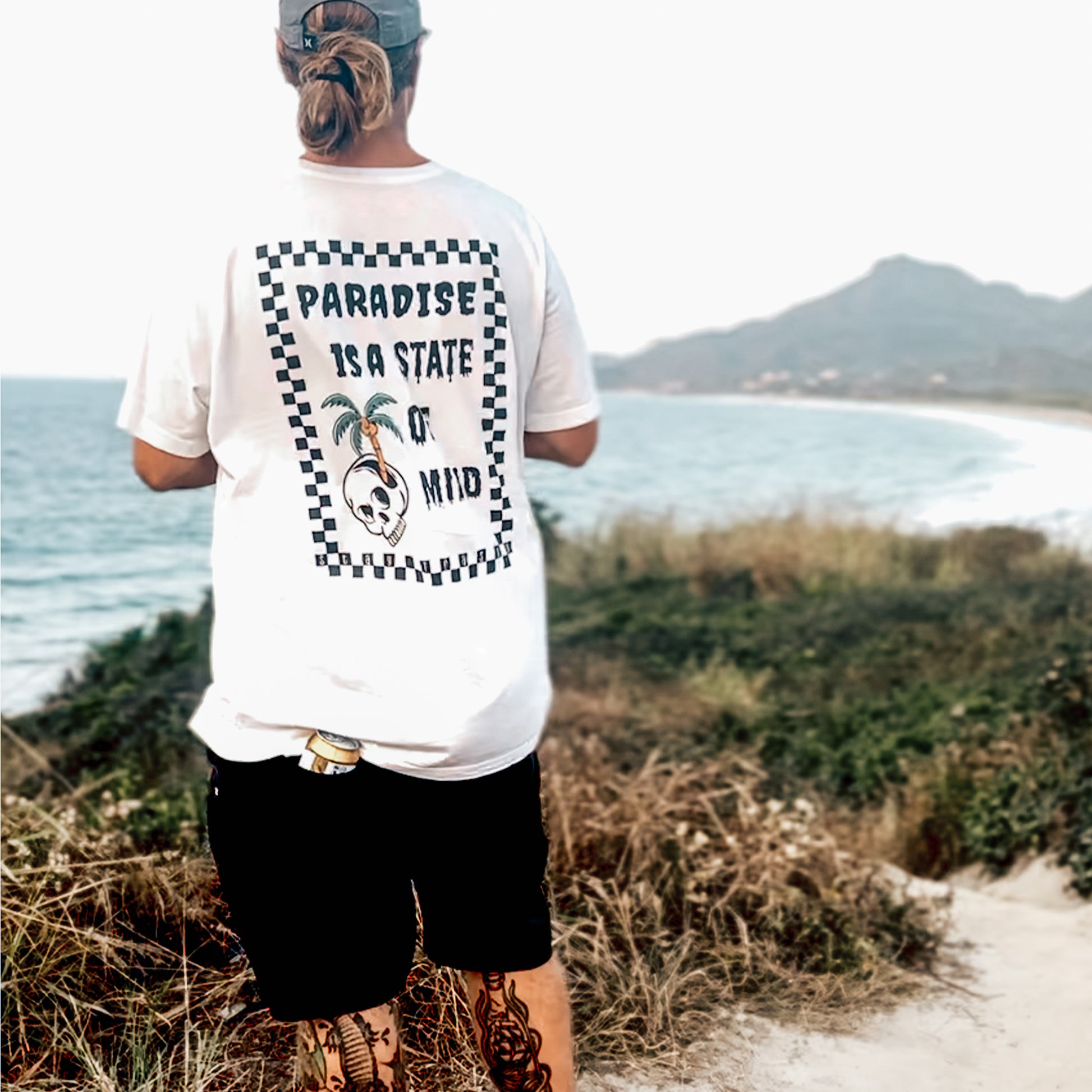 Man wearing white oversized graphic tee with 'Paradise Is A State Of Mind' back print, checkered border, palm trees and skull design, standing at beach