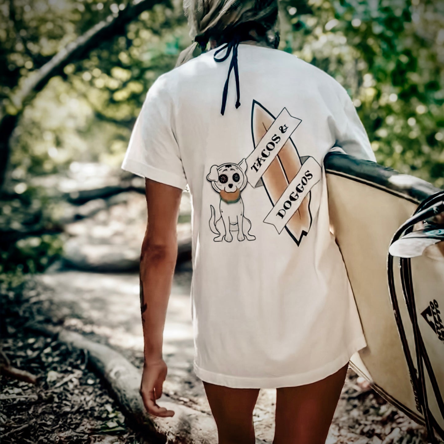 Woman carrying surfboard wearing white Tacos and Doggos tee with dog taco and surfboard back graphic, tropical path setting