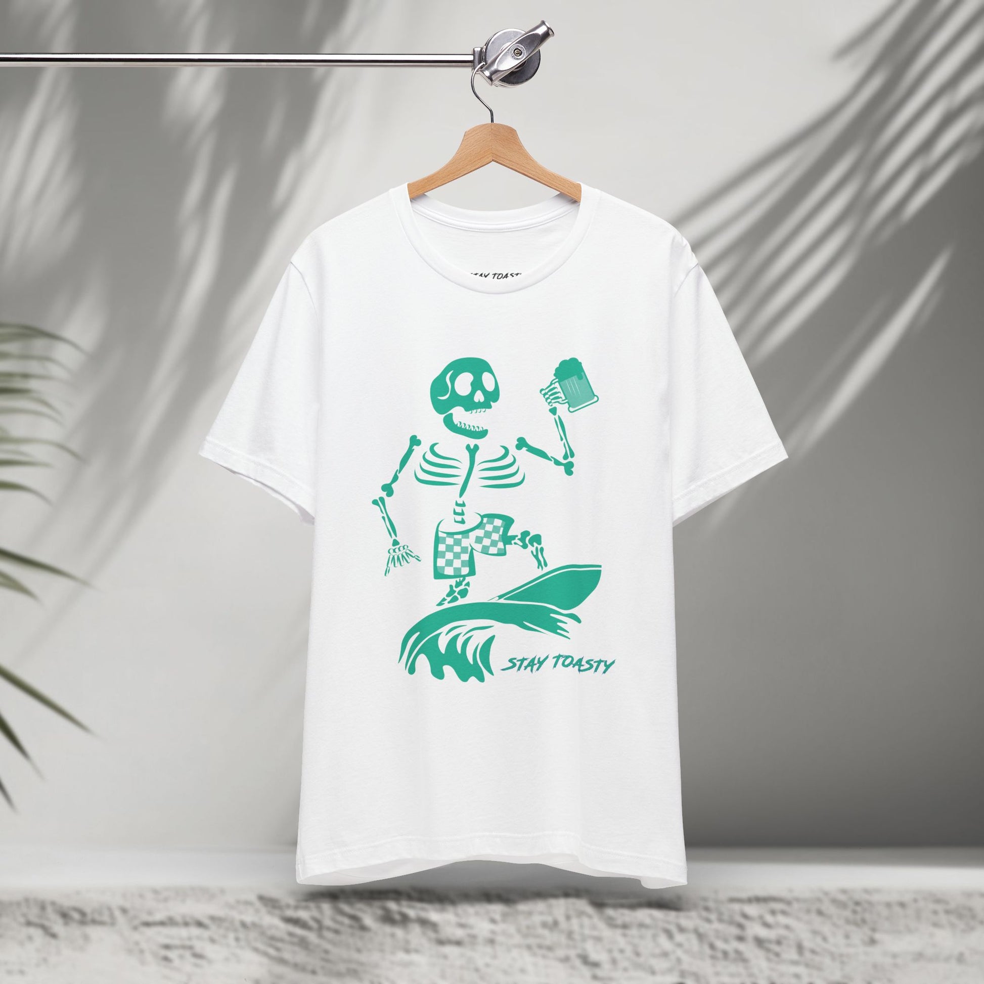 White t-shirt with a skeleton surfer design holding a beer and 'Stay Toasty' text on a hanger