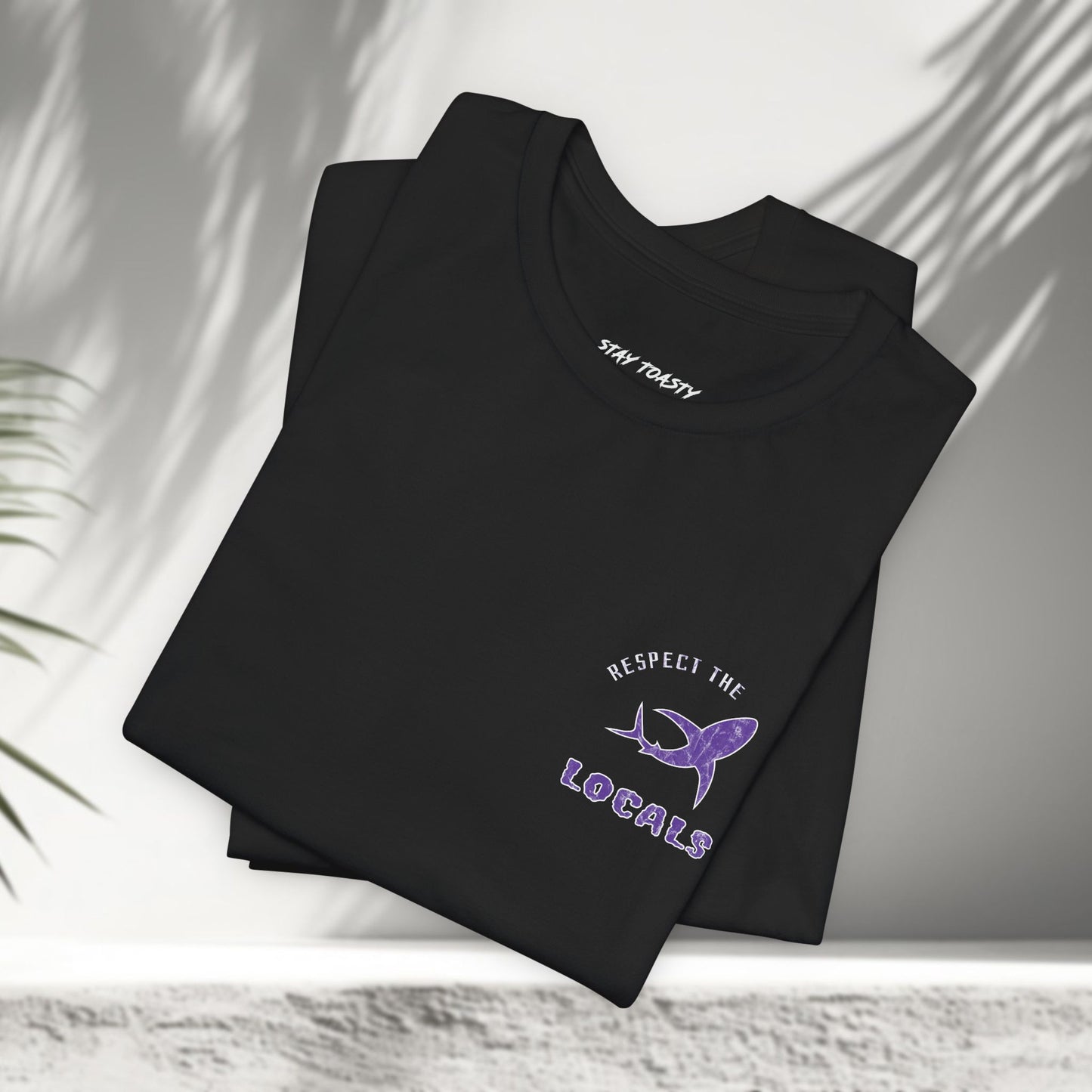 Black Respect the Locals tees folded showing purple shark graphic, tropical palm shadow background