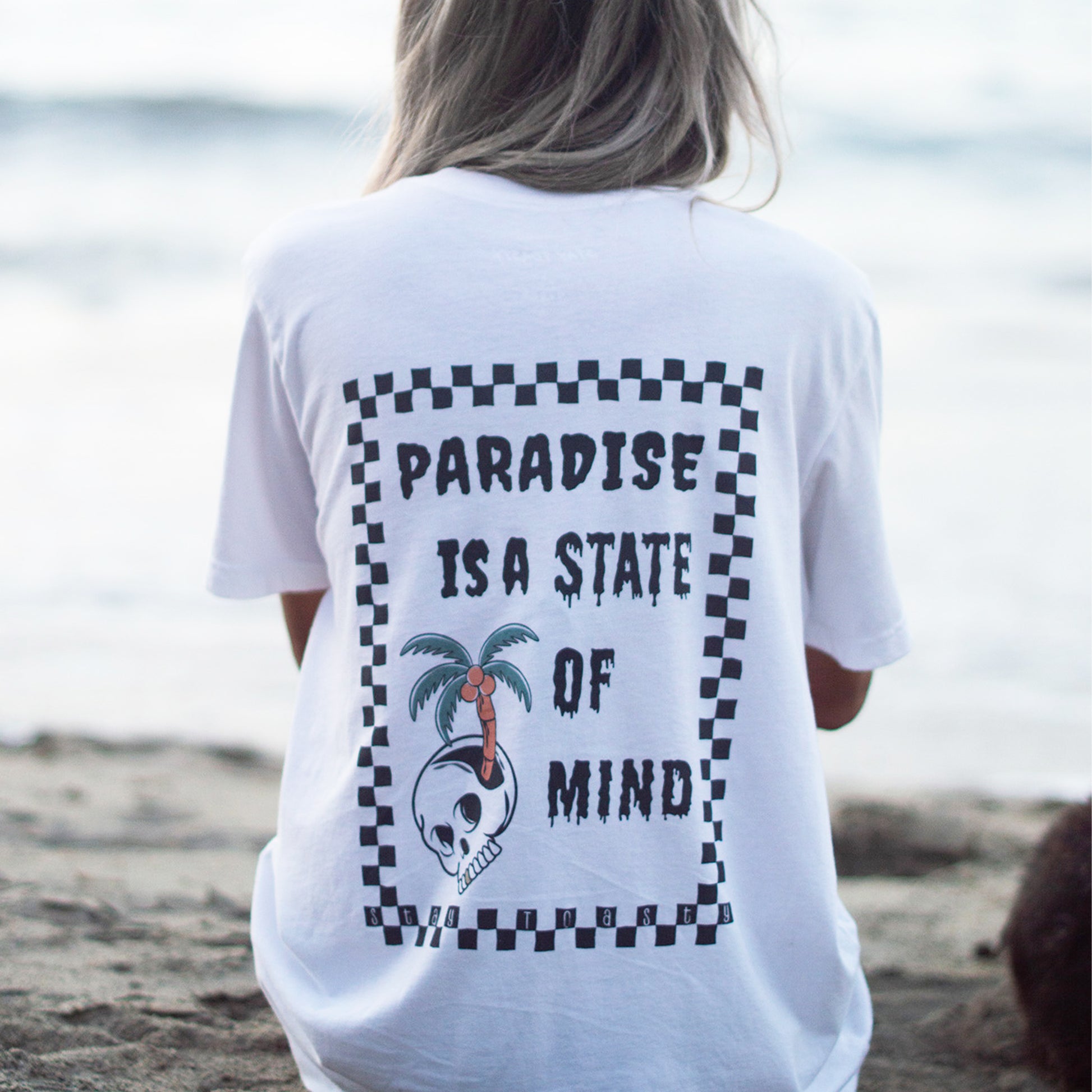 Lifestyle image of a female model wearing an oversized graphic tee. "Paradise is a State of Mind" text with a palm tree and skull design, outlined in checkers.  Shown from behind, the model is sitting on the beach looking out into the ocean. 