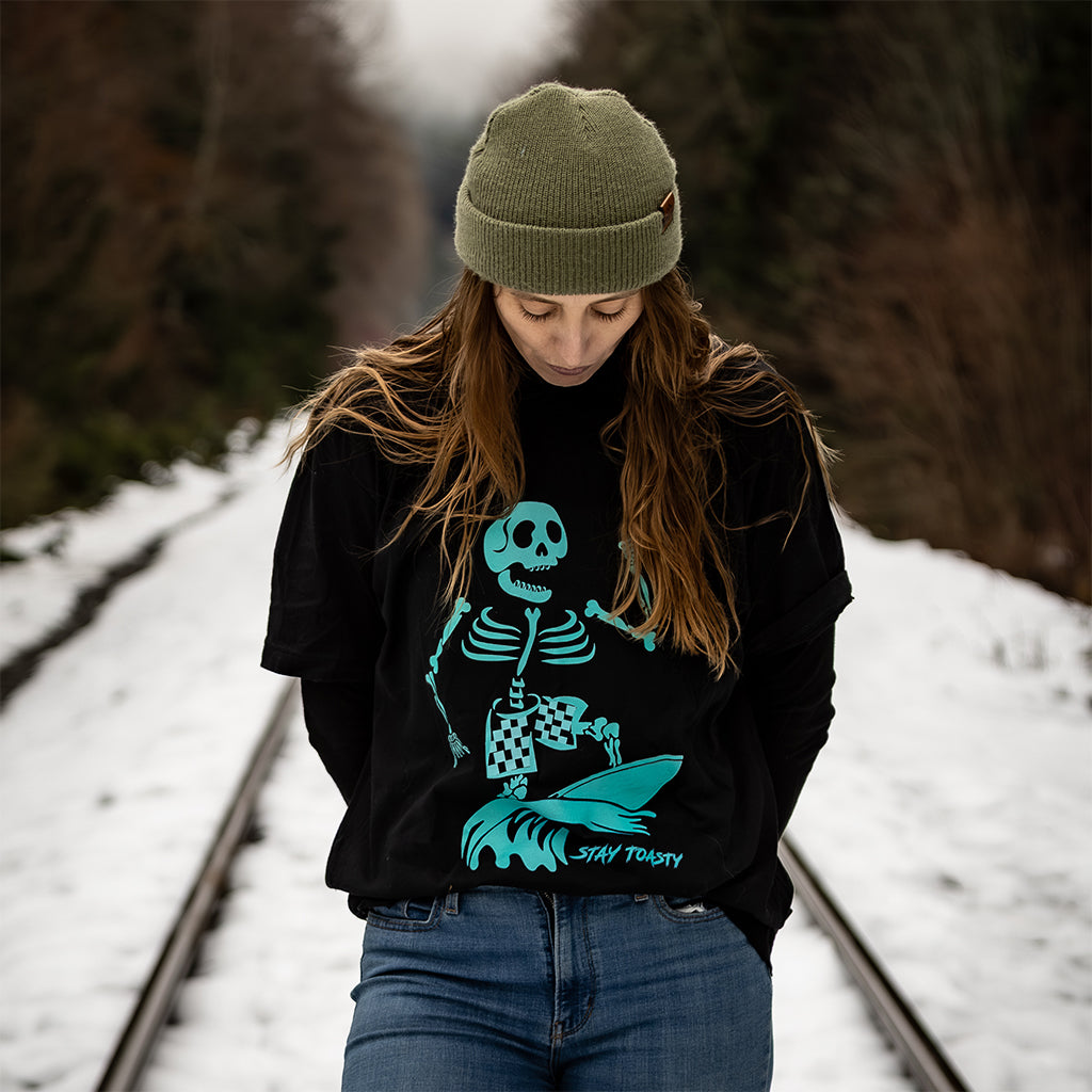 Woman in green beanie wearing black Party Wave skeleton surfer graphic tee on train tracks in snow