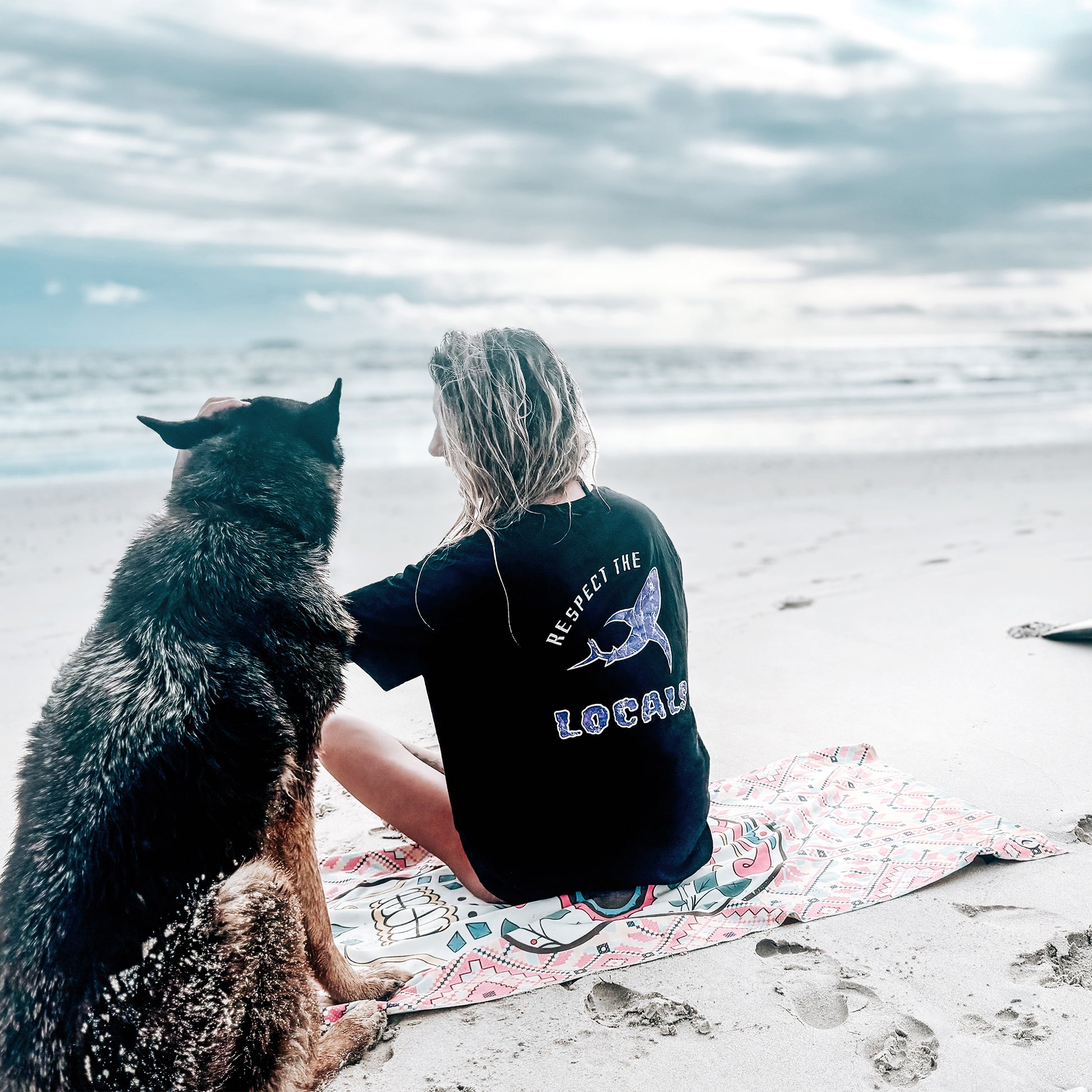 Woman sitting on beach with dog wearing black Respect the Locals tee with purple shark  back print, ocean view