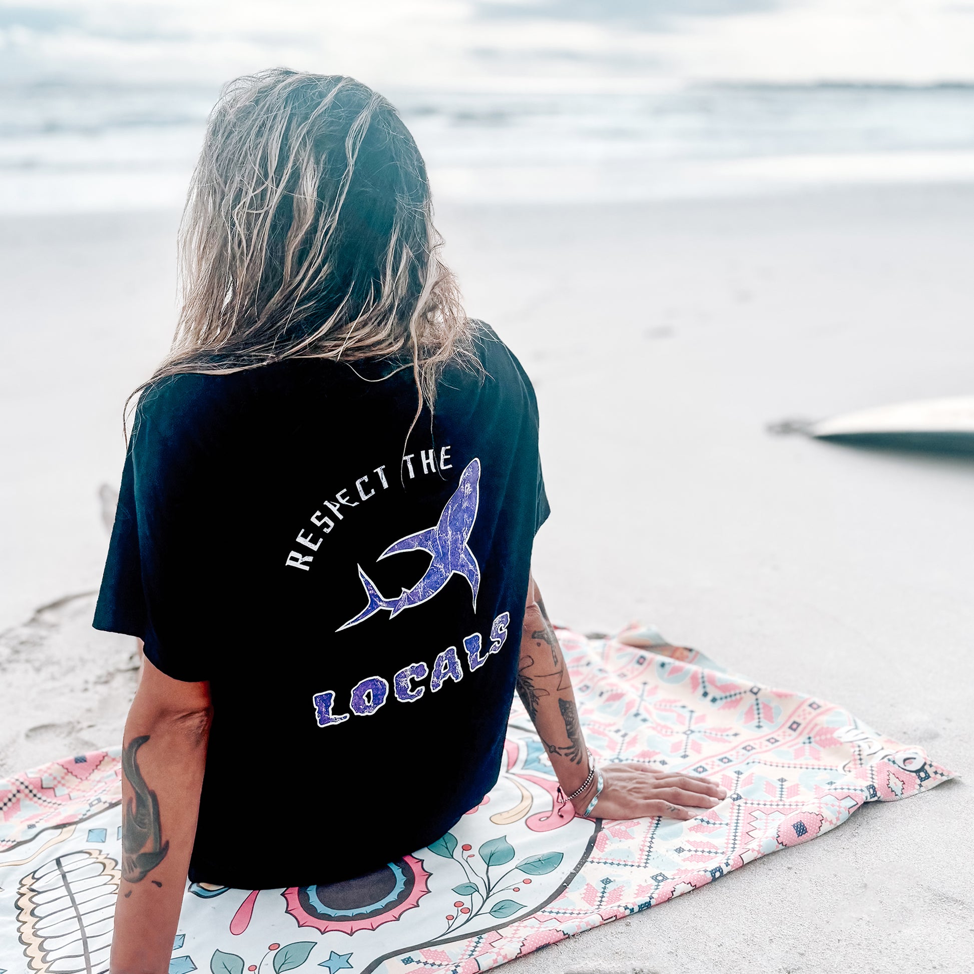 Person sitting on beach wearing black T-shirt with ‘Respect the Locals’ graphic on back.