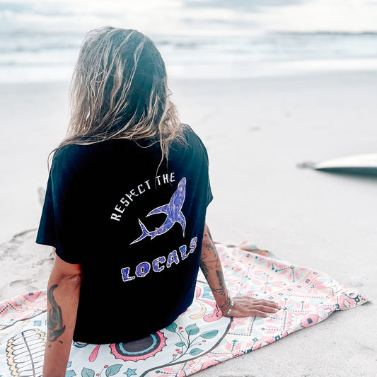 Person sitting on beach wearing black T-shirt with ‘Respect the Locals’ graphic on back.