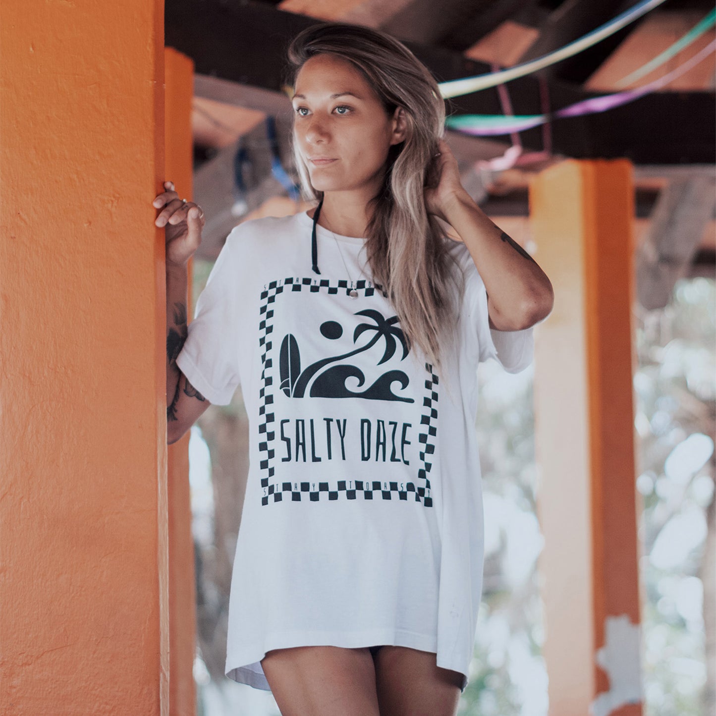 Woman standing in colourful arch in Sayulita wearing the white 'Salty Daze tee' by Stay Toasty. Tee has beach graphics and checkers. Model wearing an oversized fit. 