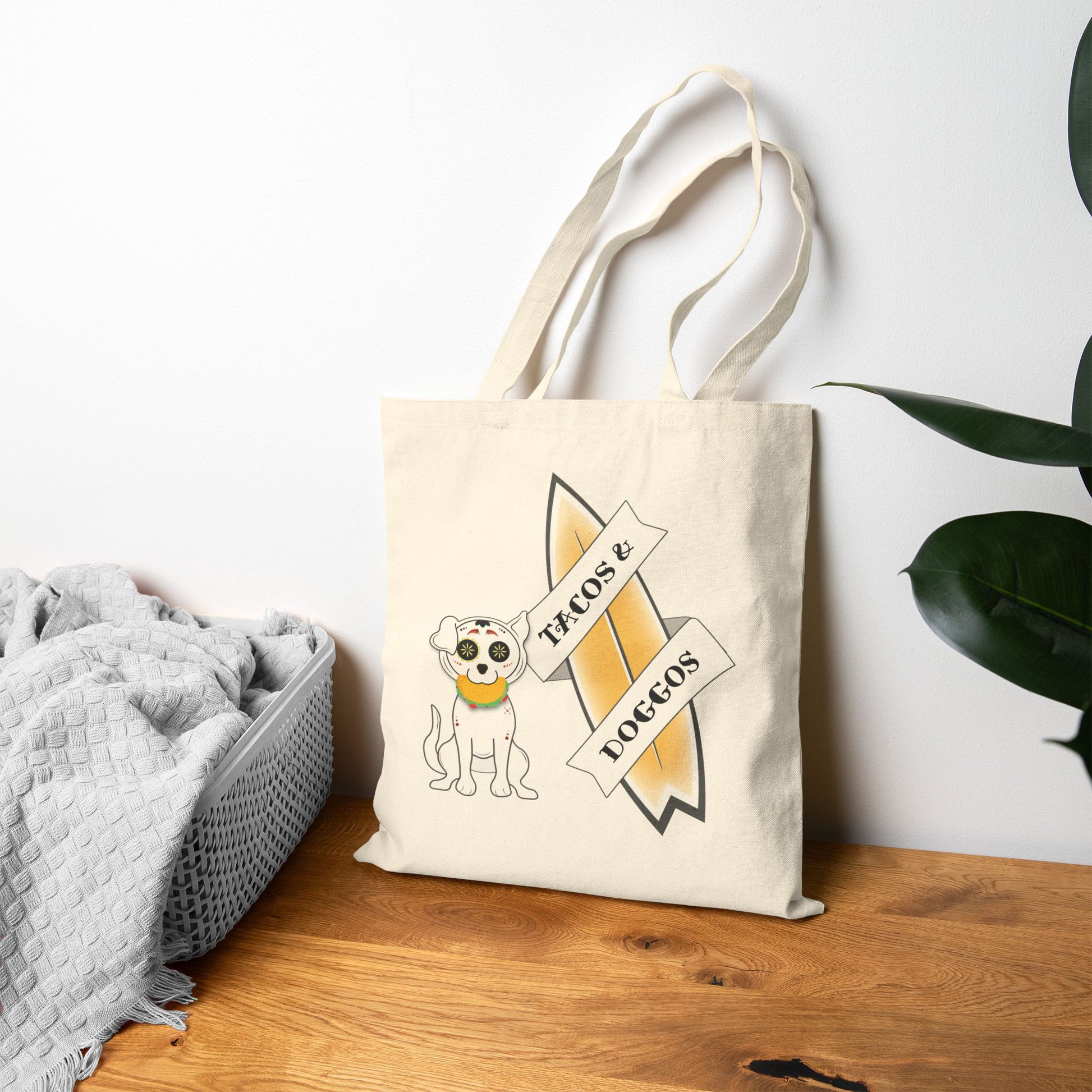 Beige tote bag with a sugar skull dog with a taco in his mouth with text 'TACOS & DOGGOS' next to a blanket and plant.
