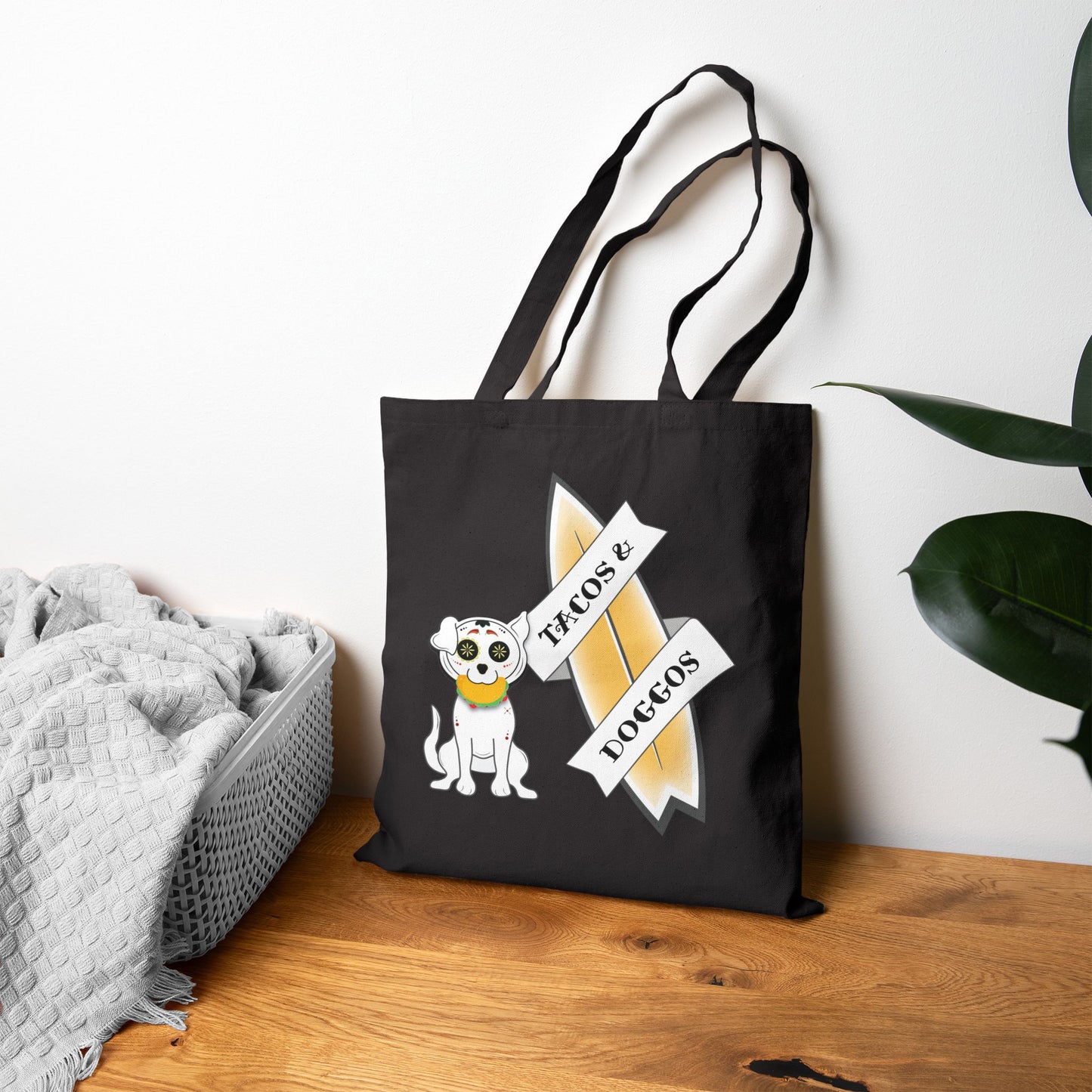 Black tote bag with a sugar skull dog with a taco in his mouth with text 'TACOS & DOGGOS' next to a blanket and plant.