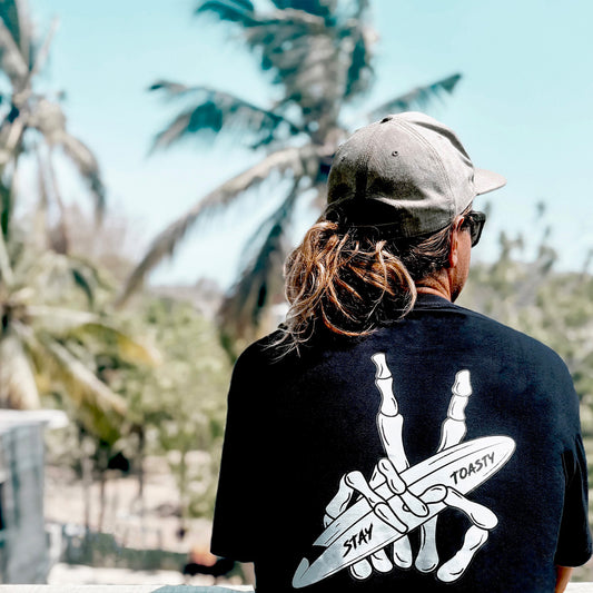Black shirt with skeleton peace sign holding surfboard, worn by person overlooking tropical scene.