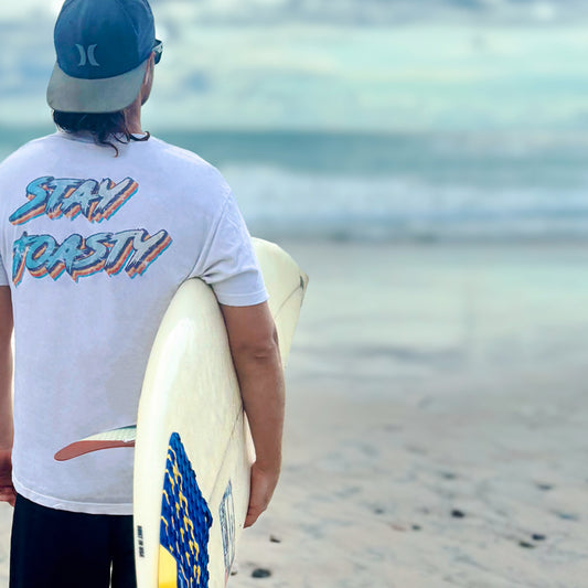 Stay Toasty’ T-shirt with vintage text worn by person holding surfboard on beach.