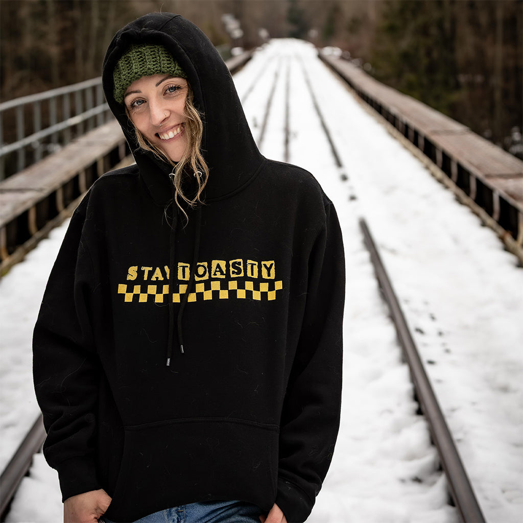 Person wearing a black hoodie with yellow 'Stay Toasty' text and checkers on snow-covered tracks