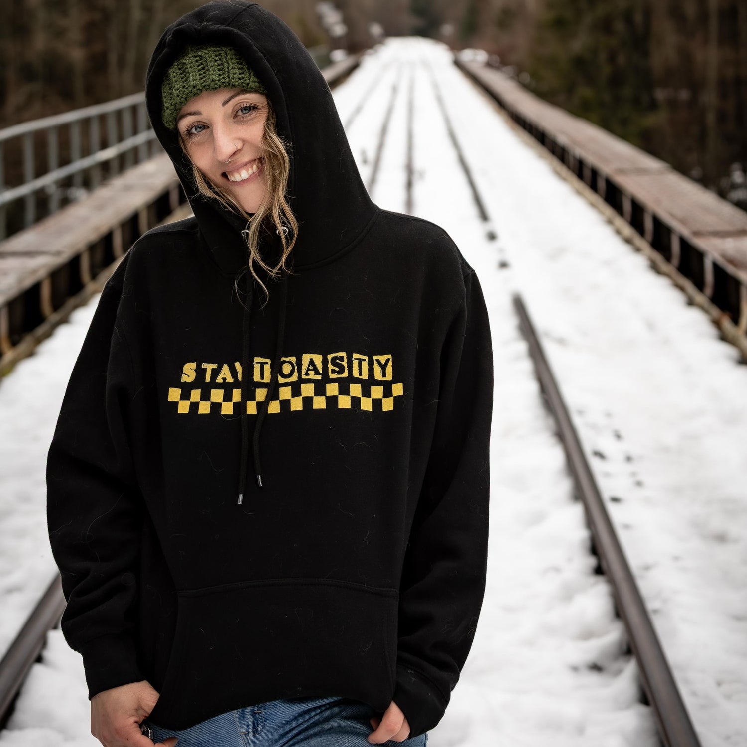 Woman wearing black Stay Toasty Wave Check Hoodie in size Large, 5'7" model, relaxed fit with hood up, snowy railway bridge Whistler BC