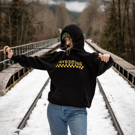 Woman wearing black Stay Toasty Wave Check Hoodie in size Large, 5'7" model, relaxed oversized fit, on snowy railway bridge in Whistler BC