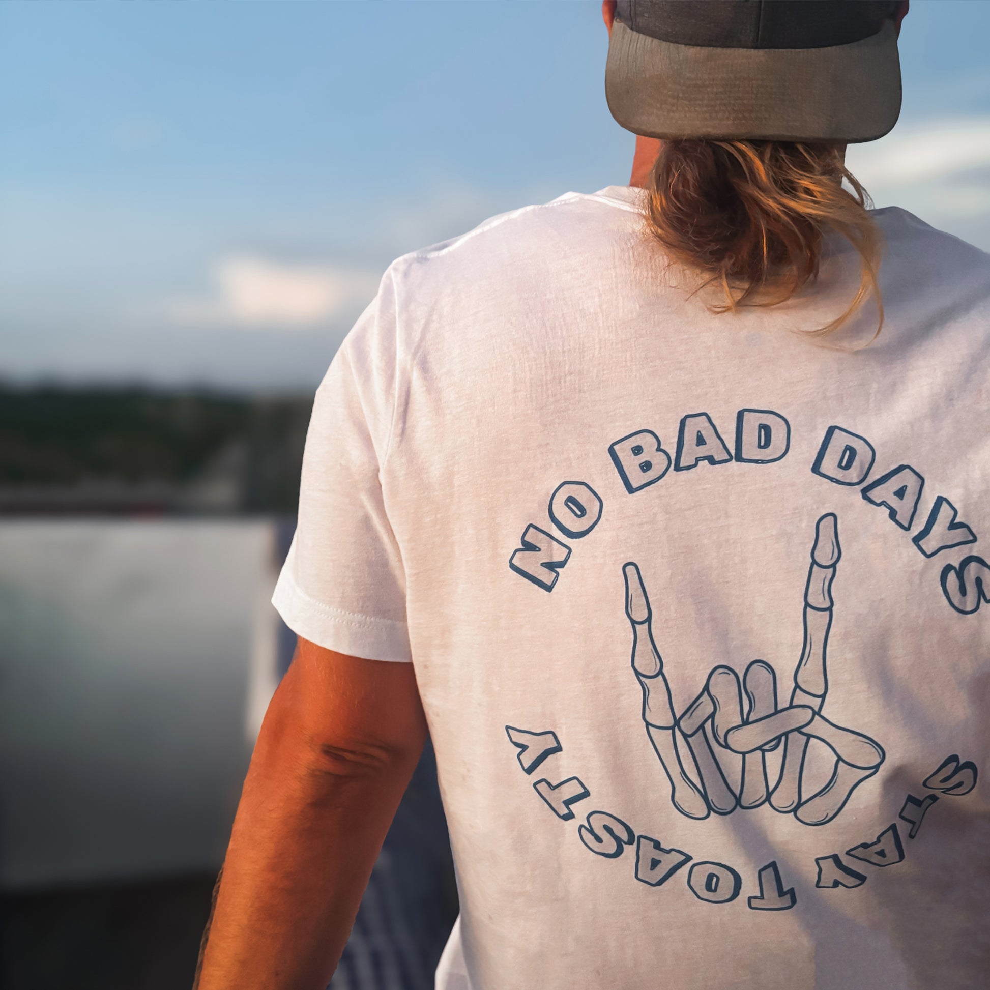 Male model sitting poolside with a white graphic tee of a skeleton hand with text "NO BAD DAYS" and "STAY TOASTY" 