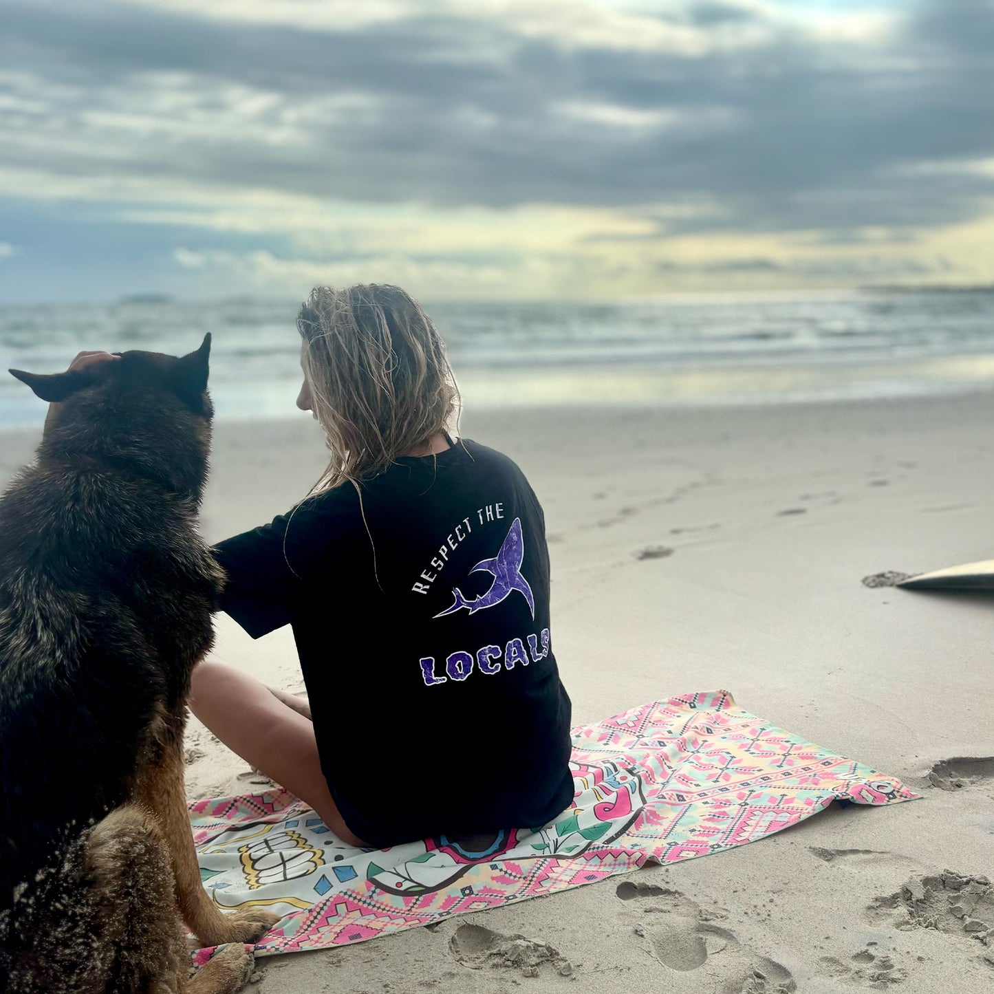 Person and dog sitting on a beach with a dog and ocean view, wearing 'Respect the Locals' t-shirt.
