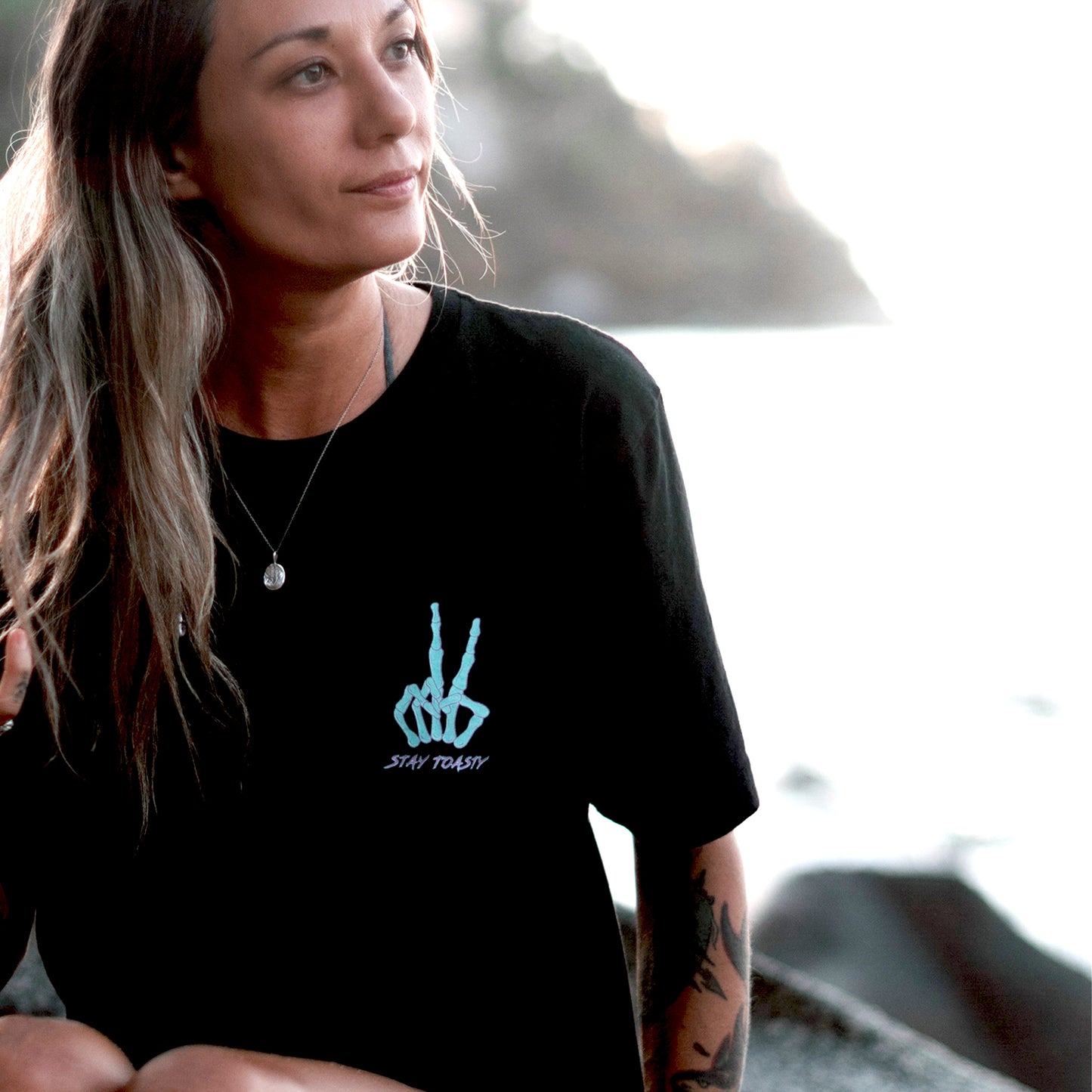 Women wearing black t-shirt with skeleton hand peace sign on front chest in mint and purple by ocean